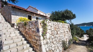 Ein Steinhaus mit rotem Ziegeldach, eine Steintreppe, die zu einer Tür führt, und im Hintergrund der Blick auf das Meer.