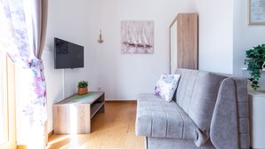 A living room with a grey sofa, a flat-screen TV, a wooden cabinet, a painting of sailboats, and a small plant. - Apartments & Rooms Cina- Standard Studio Apartment with Balcony and Sea View (Cavtat)