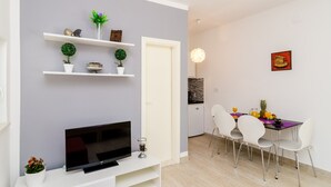Ein modernes Wohnzimmer mit einem Flachbildfernseher, weißen Regalen mit Dekoration, einem Essbereich mit Tisch und Stühlen sowie einer Küche im Hintergrund.