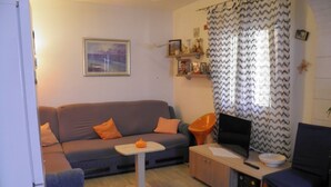 Uma sala de estar com sofá cinza, almofadas laranja, uma mesinha com brinquedo de gato, televisão, um armário de madeira e uma cadeira com almofada azul.