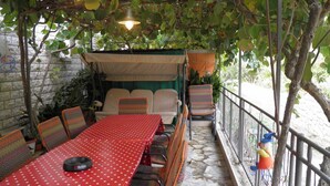 Pátio externo com mesa vermelha e cadeiras listradas, rodeado por vegetação e piso de pedra.