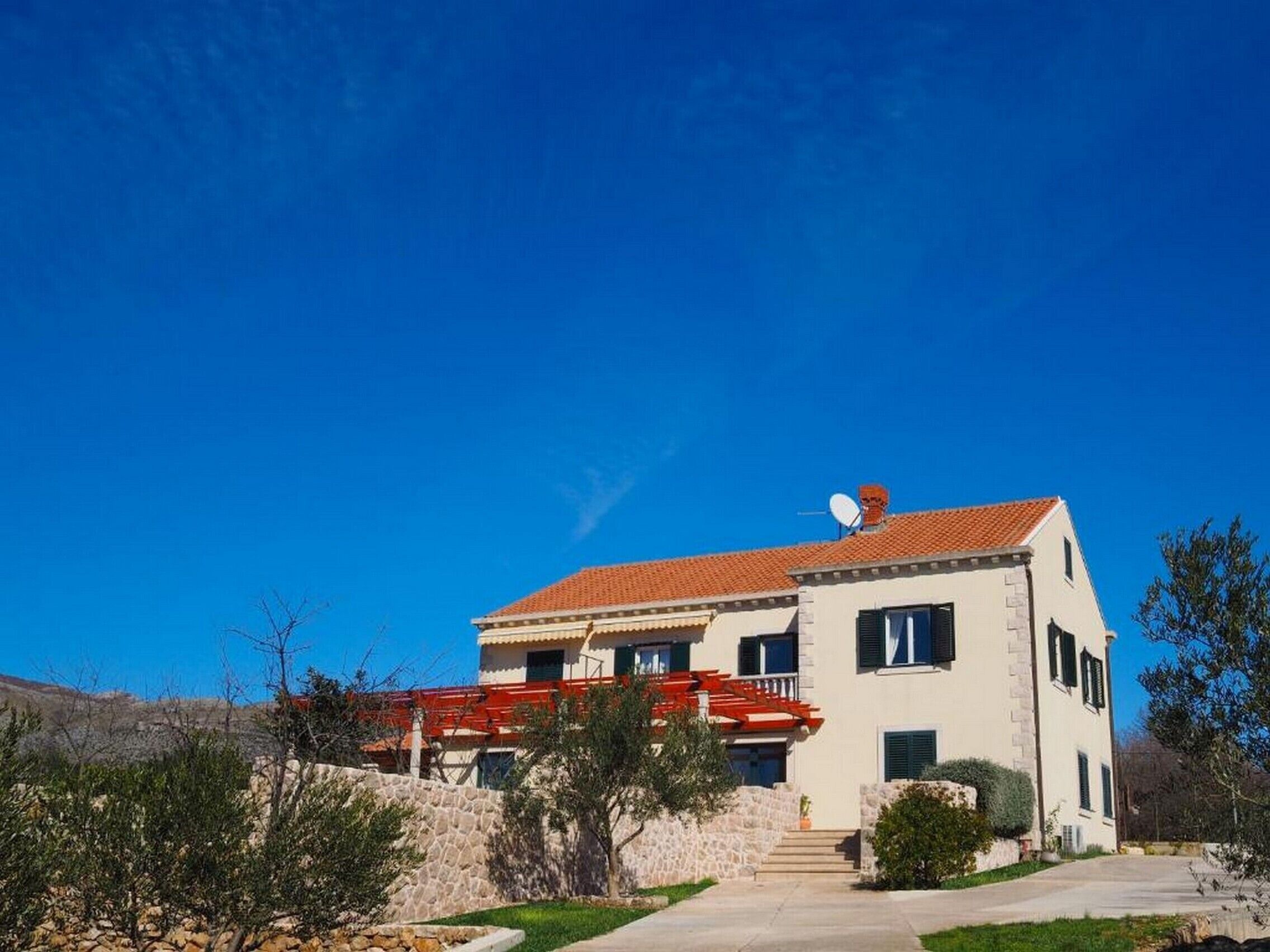 Un bâtiment à deux étages, avec un toit recouvert de tuiles rouges, une antenne parabolique et un mur de pierre en premier plan.