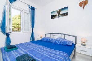 Un dormitorio con cama, ventana con cortinas, mesita de noche con lámpara y un cuadro colgado en la pared.