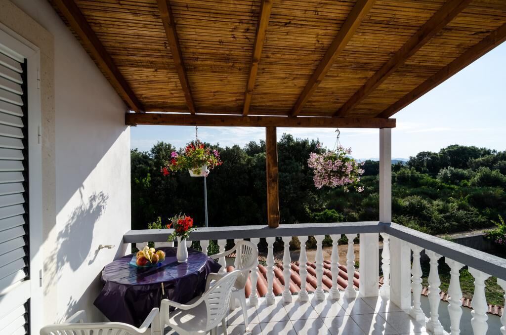 A covered balcony with a table and chairs, a view of greenery, and hanging flower baskets.