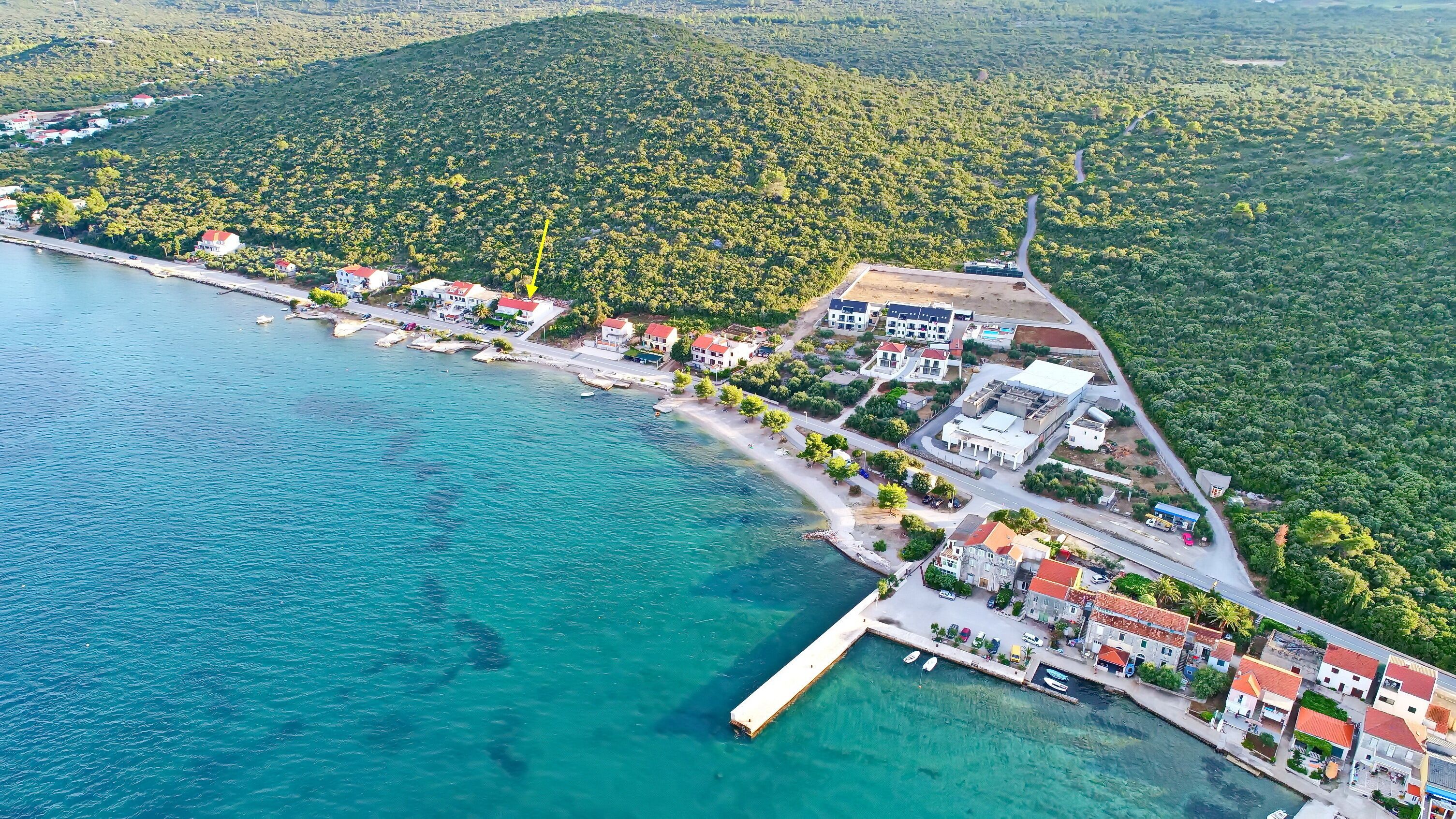 An aerial view of a coastal town with a marina, residential buildings, and a forested hillside.