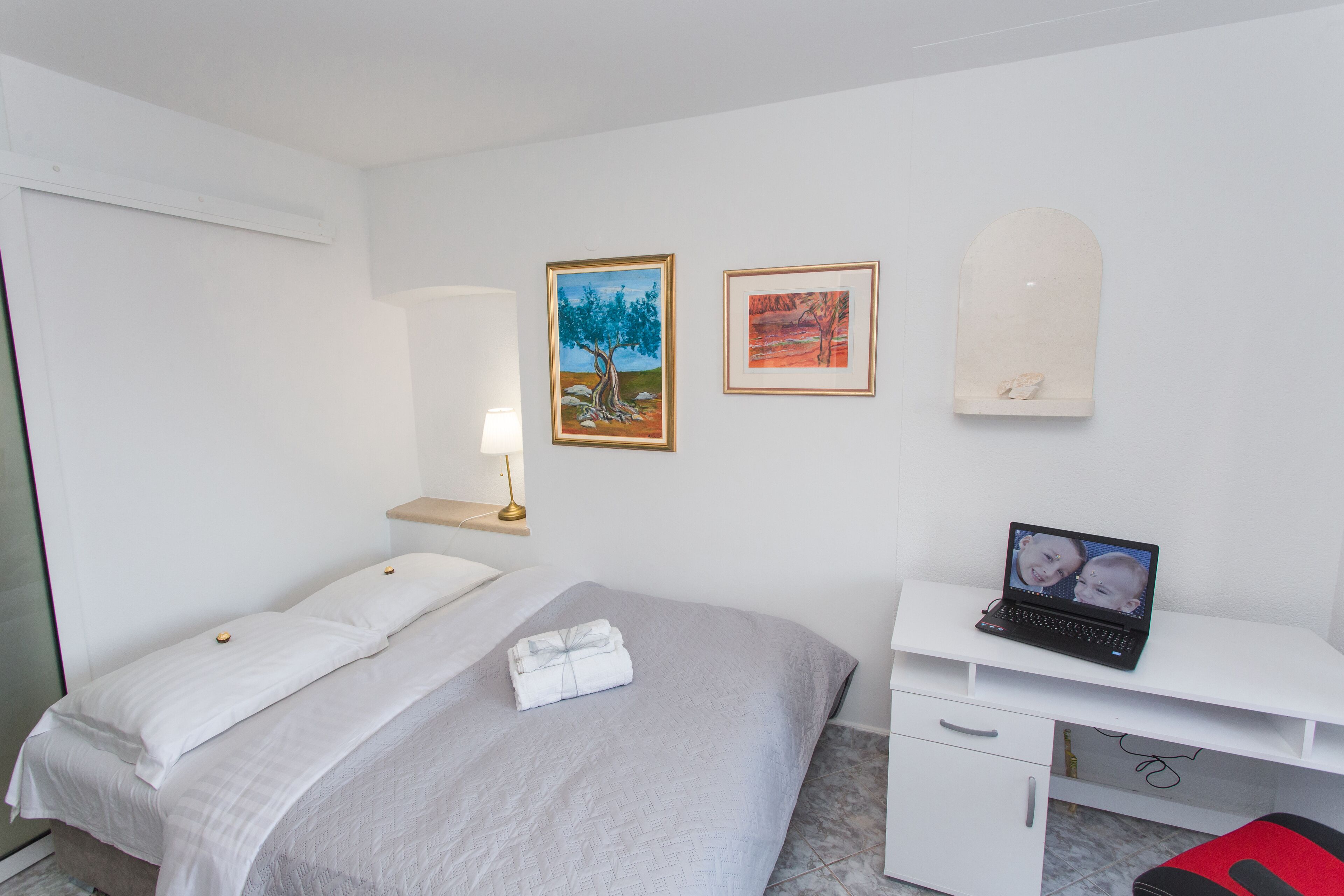 A bedroom with a bed, a desk with a laptop, a wall-mounted shelf, and two framed pictures.