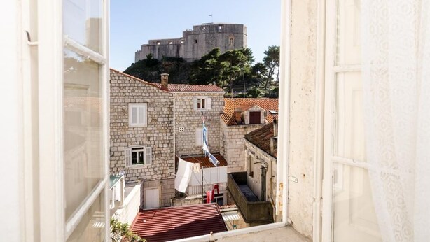 Un bâtiment en pierre, surmonté d’un toit de tuiles rouges, avec un drapeau et une vue sur une plus grande forteresse en arrière-plan.