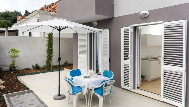 Un patio al aire libre con una mesa, sillas, un paraguas blanco y un ventanal corredizo que da a una cocina.