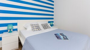 Una cama bien hecha con una colcha azul, dos almohadas y una revista sobre la cama. Hay una mesita de noche blanca con una lámpara azul turquesa junto a la cama. La pared detrás de la cama tiene rayas azules y blancas.