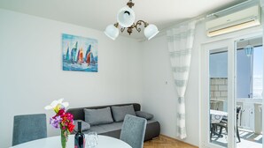 Ein modernes Wohnzimmer mit einem weißen Sofa, grauen Sesseln, einem Esstisch mit Blumenvase und einer Weinflasche, einem Gemälde von Segelbooten und einer Schiebeglastür, die zu einem Außenbereich führt.