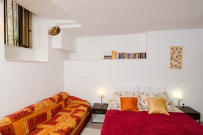 Une chambre avec un lit rouge, un canapé à motifs, une bibliothèque et une œuvre d’art murale.