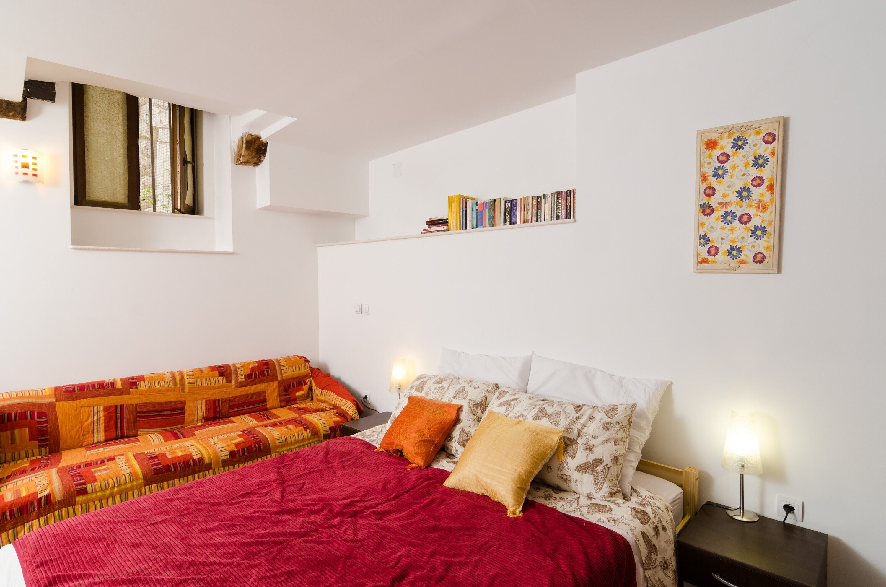Un dormitorio con cama, sofá, mesita de noche con lámpara, estante en la pared con libros y ventana con persianas.