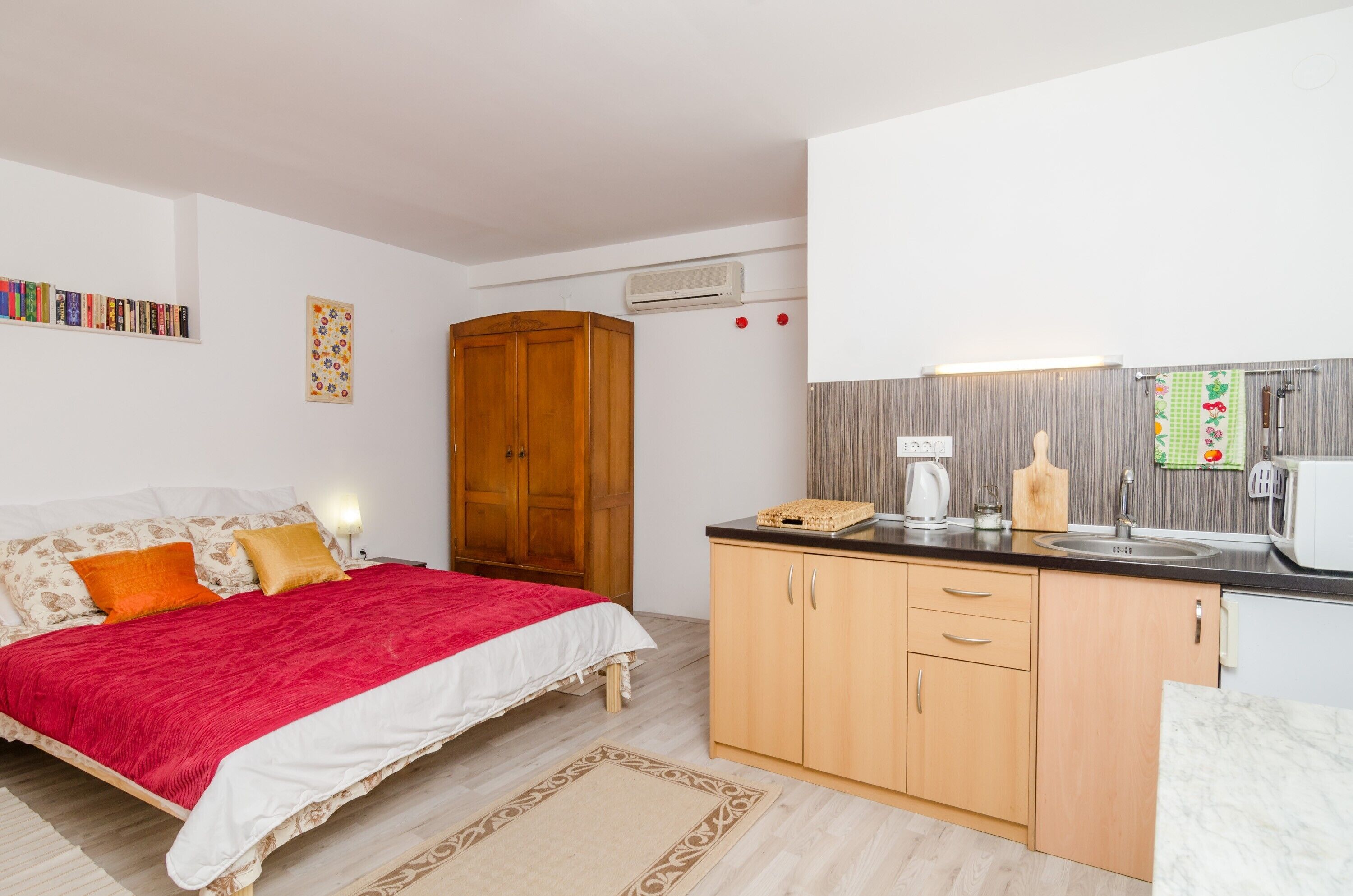 A bedroom with a bed, a nightstand, a wooden wardrobe, and a kitchen area with a sink, stove, and microwave.