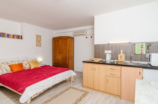 A bedroom with a bed, a nightstand, a wooden wardrobe, and a kitchen area with a sink, stove, and microwave.