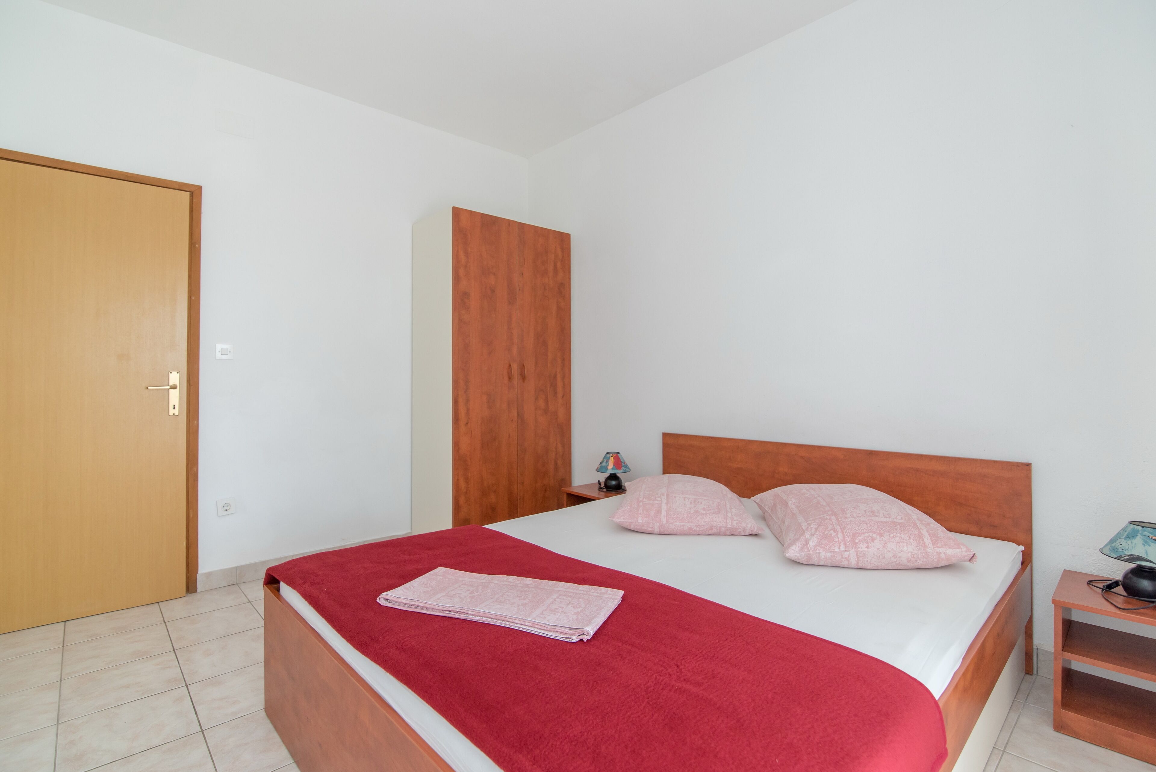 A bedroom with a wooden bed, a red blanket, and a nightstand with a lamp.