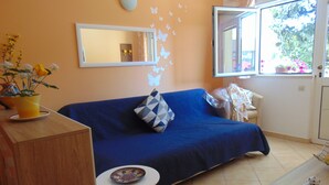 A room with a blue sofa, a small white table, a wooden cabinet, a mirror, and a window with a view of greenery. - Apartments Elelu-One-Bedroom Apartment with Terrace (Dubrovnik)