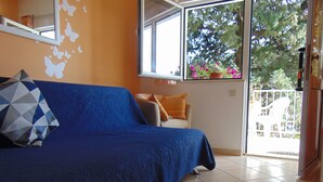 A room with a blue sofa, a patterned cushion, a small table with a decorative plate, a colorful rug, and a window with a view of trees and a balcony. - Apartments Elelu-One-Bedroom Apartment with Terrace (Dubrovnik)