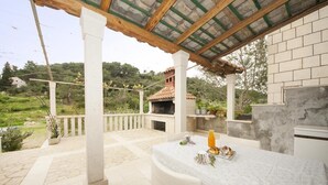 A covered outdoor dining area with a table set for two, a brick oven, and a view of a green landscape. - Apartments Kalajzic- One-Bedroom Apartment with Terrace (Pucisca)