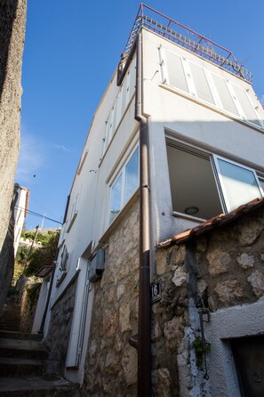 Un immeuble de deux étages doté d’une façade en pierre, d’un balcon en métal et d’une fenêtre visible avec des volets.