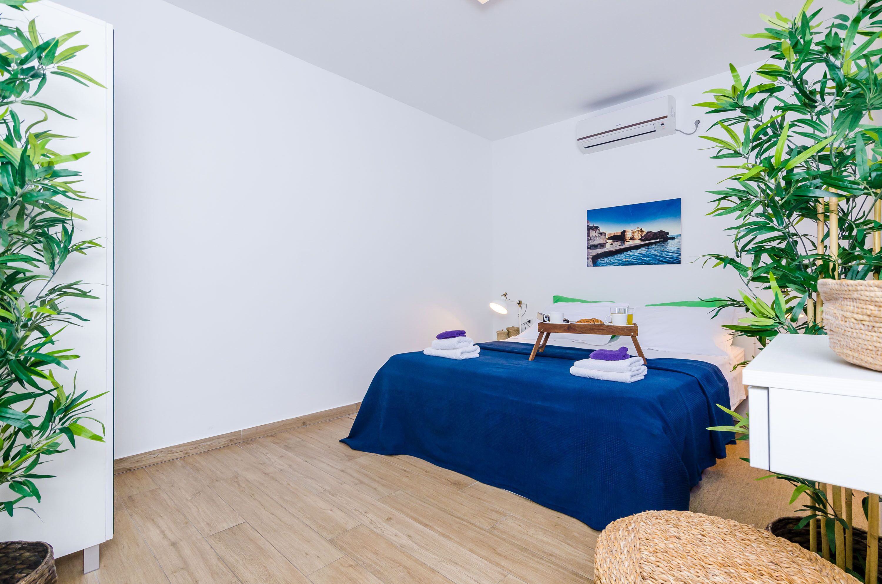 A room with a bed, a small table with towels, a picture on the wall, and potted plants.