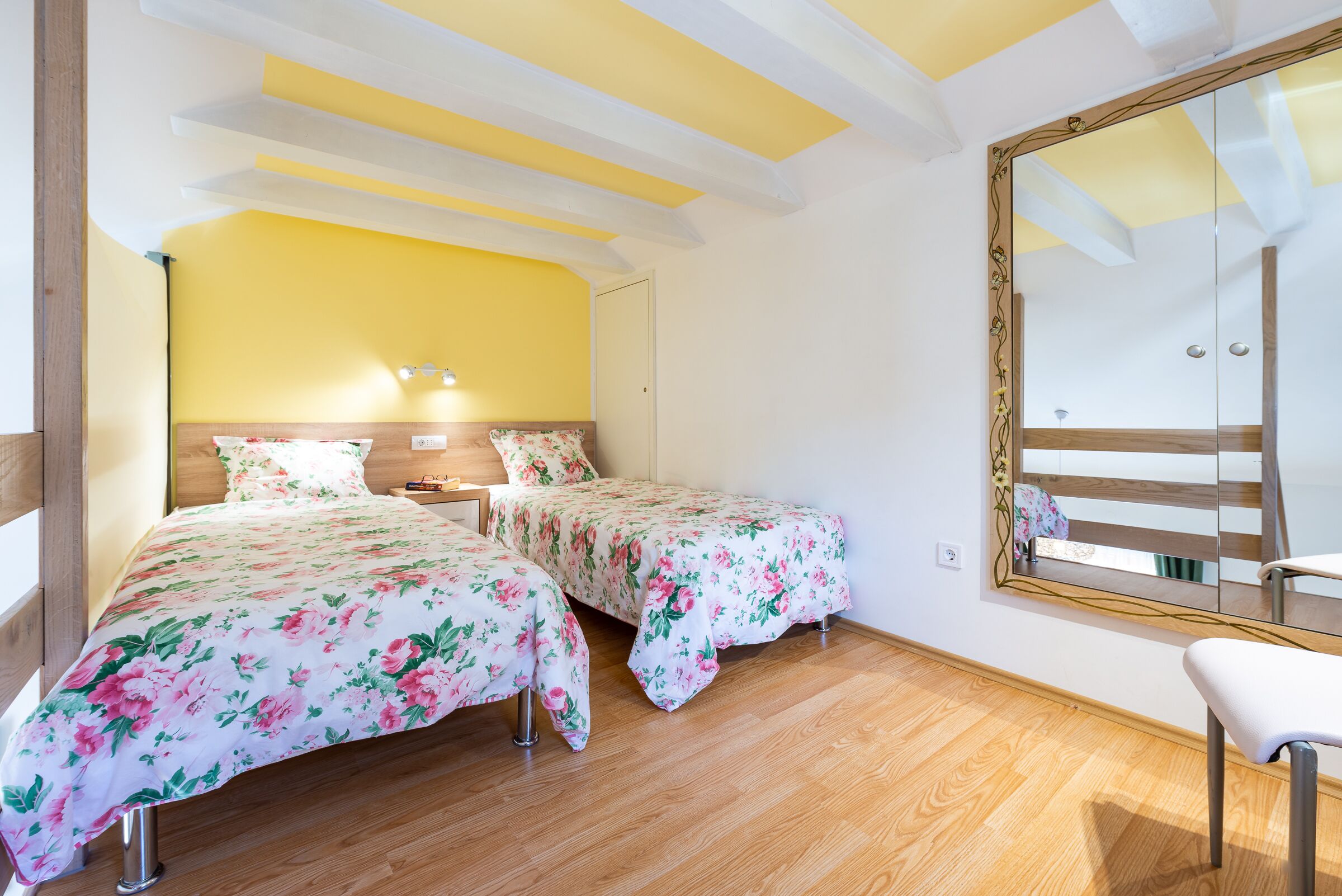 A room with two beds, wooden flooring, a large mirror, and a bench.