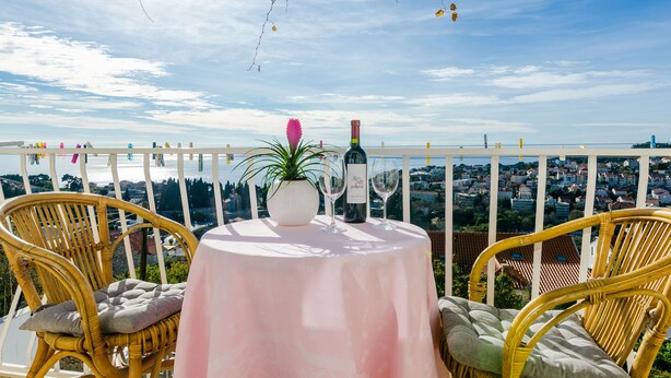 A balcony with wicker chairs, a table with a white tablecloth, a bottle of wine, and a vase with a flower. - Rooms Batina- Standard Double Room (Dubrovnik)