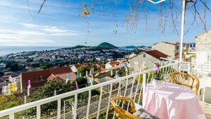 A balcony with a table and chairs, overlooking a coastal town and a mountain. - Rooms Batina- Standard Double Room (Dubrovnik)