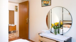 Un dormitorio con puerta de madera, un espejo redondo, una mesita de noche con cubierta de encaje blanco y una cama con manta blanca.