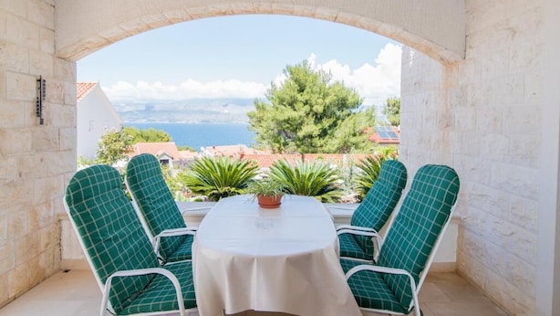 A balcony with two green checkered lounge chairs and a table, overlooking a scenic view of the sea and buildings.