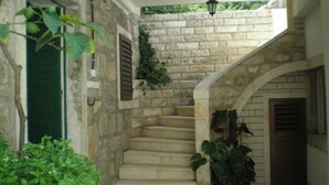 Un escalier en pierre mène à une porte ornée d’un volet vert, le tout étant entouré de verdure et surplombé d’un luminaire suspendu.