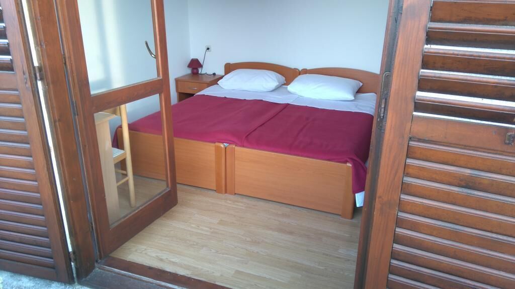 Een houten bed met een rode deken, een nachtkastje met een lamp en een houten deur.