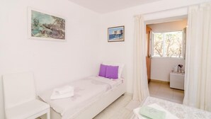 Ein kleines, gut beleuchtetes Schlafzimmer mit einem Einzelbett, einem Sessel und einem kleinen Tisch. An der Wand hängen zwei gerahmte Bilder und es gibt ein Fenster mit Vorhängen.