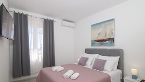 A neatly made bed with a pink bedspread, a painting of a sailboat on the wall, and a white nightstand with a lamp.
