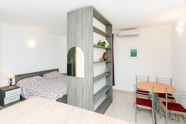 A bedroom with a bed, a nightstand, a desk, a chair, a table, and a shelf unit.