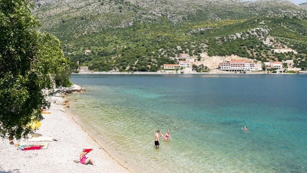Escena costera con aguas transparentes, una playa arenosa y edificios en una colina.