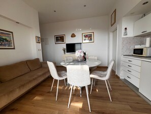 A modern living space with a round dining table, four chairs, a sofa, a kitchen area with a microwave, and framed artwork on the walls.