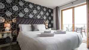 A hotel room with a large bed, a dark tufted headboard, a floral wallpaper, and a balcony with outdoor furniture. - Penthouse B (Newquay)