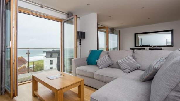 A modern living room with a grey sofa, a wooden coffee table, and a large sliding glass door leading to a balcony with a view of the sea. - Penthouse B (Newquay)