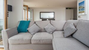 A modern living room with a grey sectional sofa, a teal throw blanket, and striped pillows. A flat-screen TV is mounted on the wall above the sofa. - Penthouse B (Newquay)