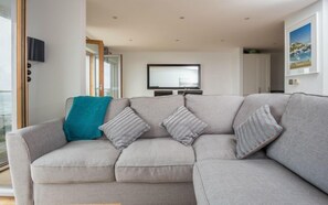 A modern living room with a grey sectional sofa, a teal throw blanket, and striped pillows. A flat-screen TV is mounted on the wall above the sofa.