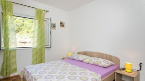 Un dormitorio con cama, mesita de noche, ventana con cortinas y un pequeño cuadro en la pared.