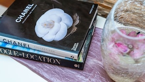 Une pile de livres, dont « Chanel Collections et Créations » et « Vogue en tournage », est posée sur une surface texturée, à côté d’un vase en verre décoratif contenant des fleurs.