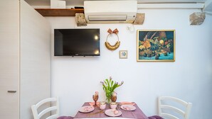 Un coin repas avec une table dressée pour quatre personnes, un téléviseur à écran plat fixé au mur et un tableau représentant des fruits et des feuilles.