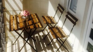 Ein kleiner Außenbereich mit Tisch und zwei Stühlen, einer Vase mit Blumen und einem Fenster mit Jalousien.