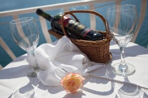 Una mesa con una botella de vino, dos copas de vino, una rosa y un mantel blanco, en un día soleado junto al agua.