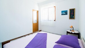 A room with a bed, a door, a window with curtains, a small table with a lamp, and two framed pictures on the wall. - Apartments Veve - One Bedroom Apartment with Terrace Sea View (Postira)