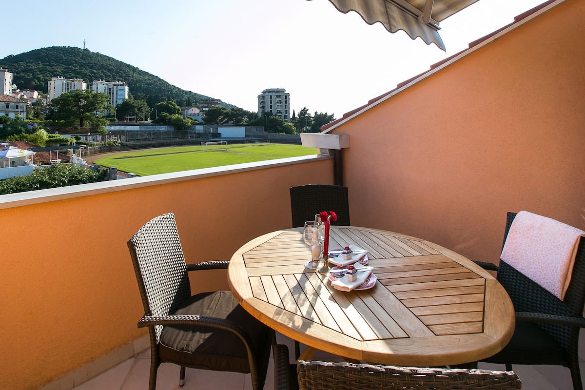 A balcony with a round wooden table, two chairs, and a small table setting with a glass, a red napkin, and a plate with a cake. A view of a green field and buildings is visible in the background.