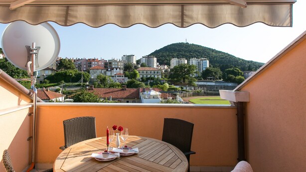 Een balkon met een ronde houten tafel gedekt voor twee personen, een schotelantenne en een uitzicht op een stadsgezicht met gebouwen en een heuvel.