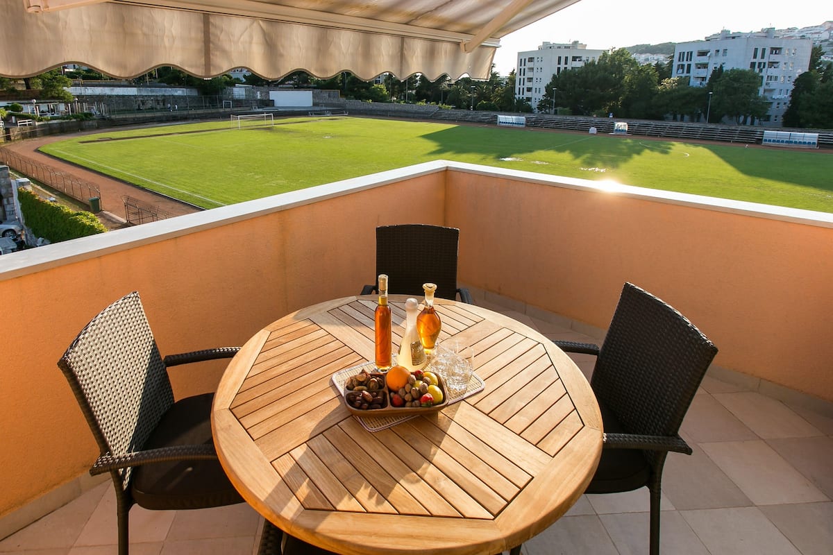 A round wooden table with two chairs, a bottle of wine, and a glass on the balcony overlooking a soccer field.