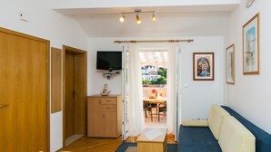 Een knus woongedeelte met een houten garderobe, een aan de muur bevestigde televisie, een bank, een eettafel met stoelen, en uitzicht op buiten via een deuropening.